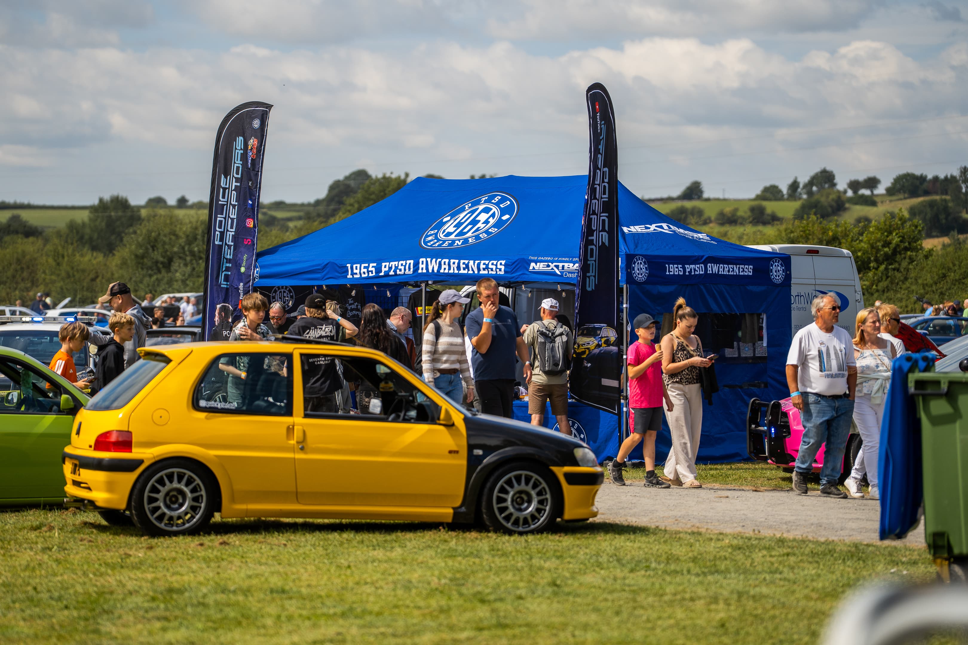 Visitors browsing stalls and a yellow modified hatchback at a WUN meet, with community gazebos in the background